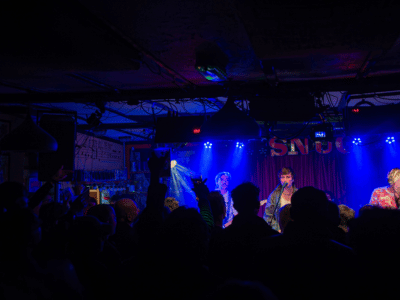 Live band performing at The Snug, with the stage lit in blue and purple and a crowd gathered in front of the performers.